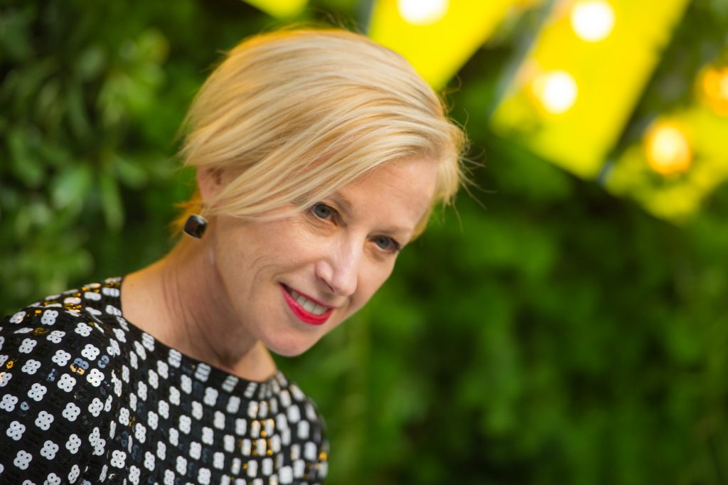 A photograph of Cindy Sherman from the neck up in a blonde bob and checkered dress before a green and yelow background