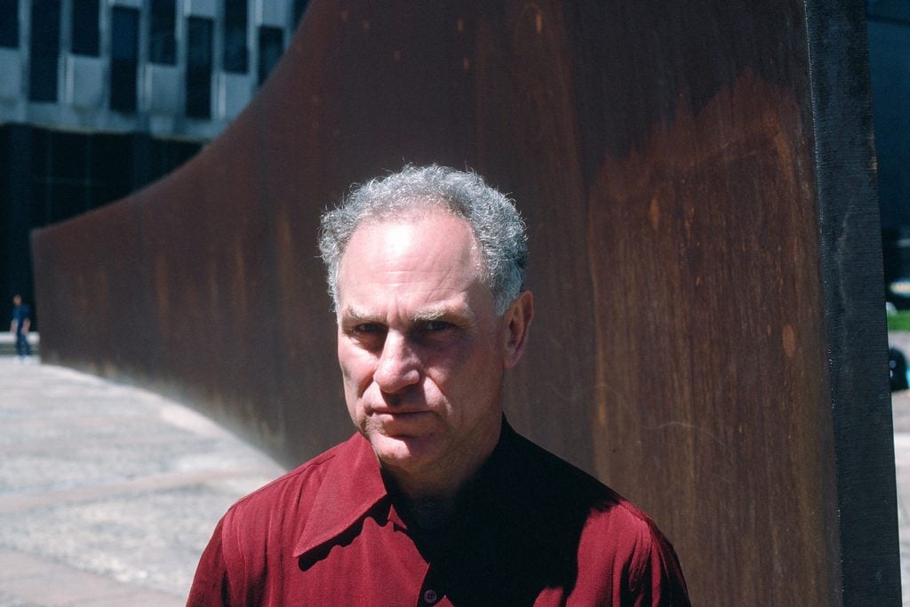 Richard Serra wears a red shirt and stands in front of his work