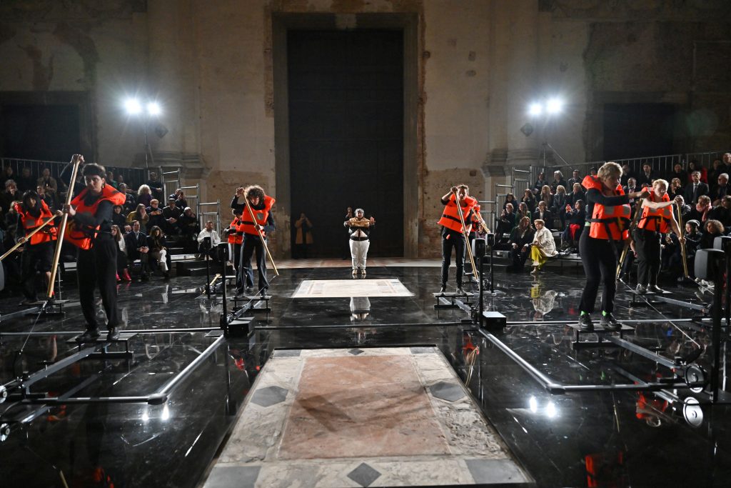 a group of artists wearing orange life vests performs an artwork in a deconsecrated church in venice
