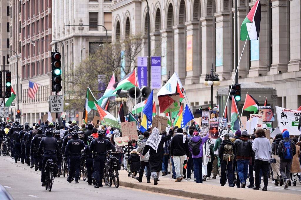 The image shows a group of people marching in the street in downtown Chicago, possibly as part of a protest. The crowd includes individuals carrying Palestine flags and a line of police in riot gear line the street.