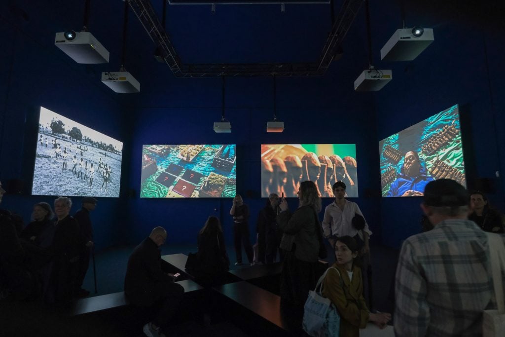 The image shows a group of people standing in front of a large screen. The setting appears to be indoors, possibly at an exhibition related to art.