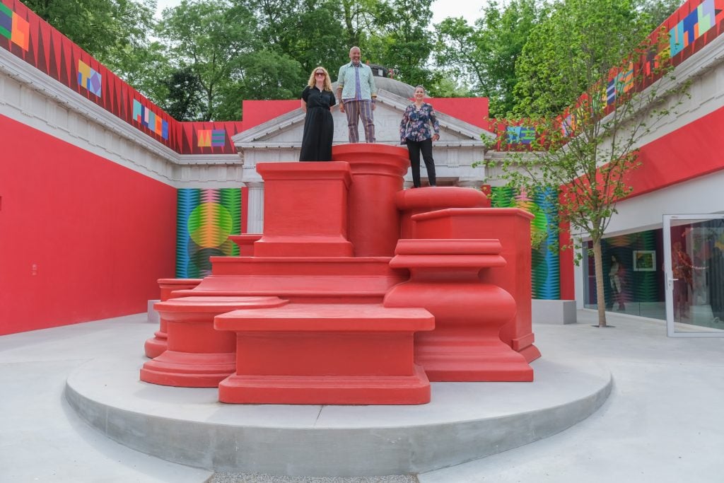 The image shows a group of people standing on a red staircase outdoors. There are trees and a street visible in the background. The setting seems to be related to the Jeffrey Gibson US Pavilion at the Venice Biennale.