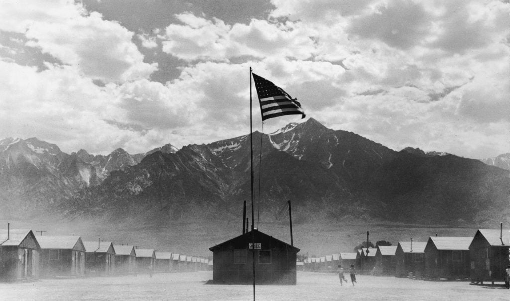 A black and white photograph of an American flag at the center of an incarceration camp at the foot of the Sierra Nevada mountains