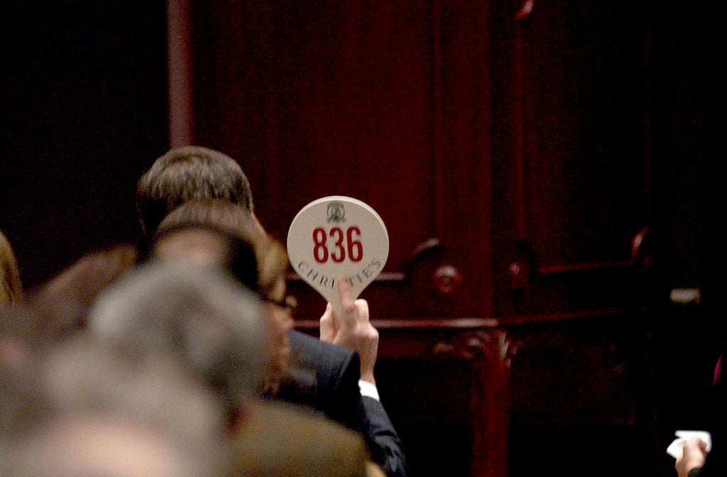 In a color photo, people's heads are seen from the back, sitting in a dark room. One person raises a white paddle with the number 836 on it.