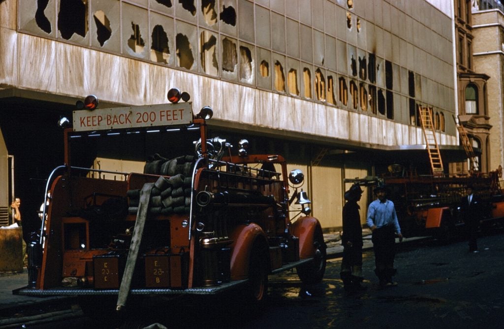 A brilliant film photograph of a 1950s firetruck in front of a fire singed MoMA with blackened, broken windows