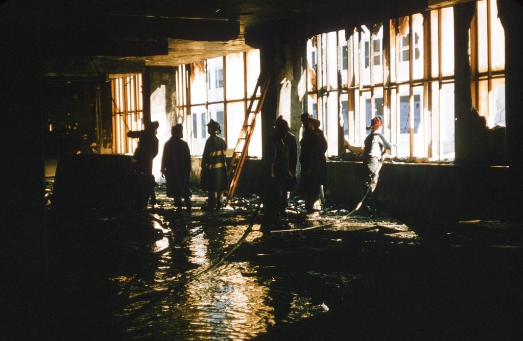 A photograph of a dark, dilapidated room with inches of water visble on the floor and bright sunlights pouring in through busted out window panes