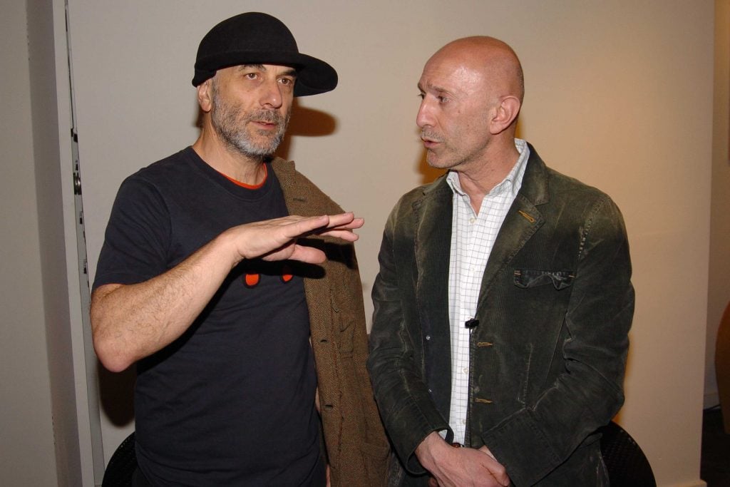 The image shows two men engaged in what appears to be a serious conversation. The man on the left is wearing a black t-shirt with an orange design on the front and a dark hat, gesturing with his hand as if explaining something. The man on the right, who is listening attentively, is wearing a white checkered shirt and a dark green corduroy jacket. They both appear to be middle-aged, and the setting looks like an indoor space, possibly a gallery or at an informal event. The context suggests they might be discussing something related to the event or sharing insights on a common interest. Is this conversation helpful so far?
