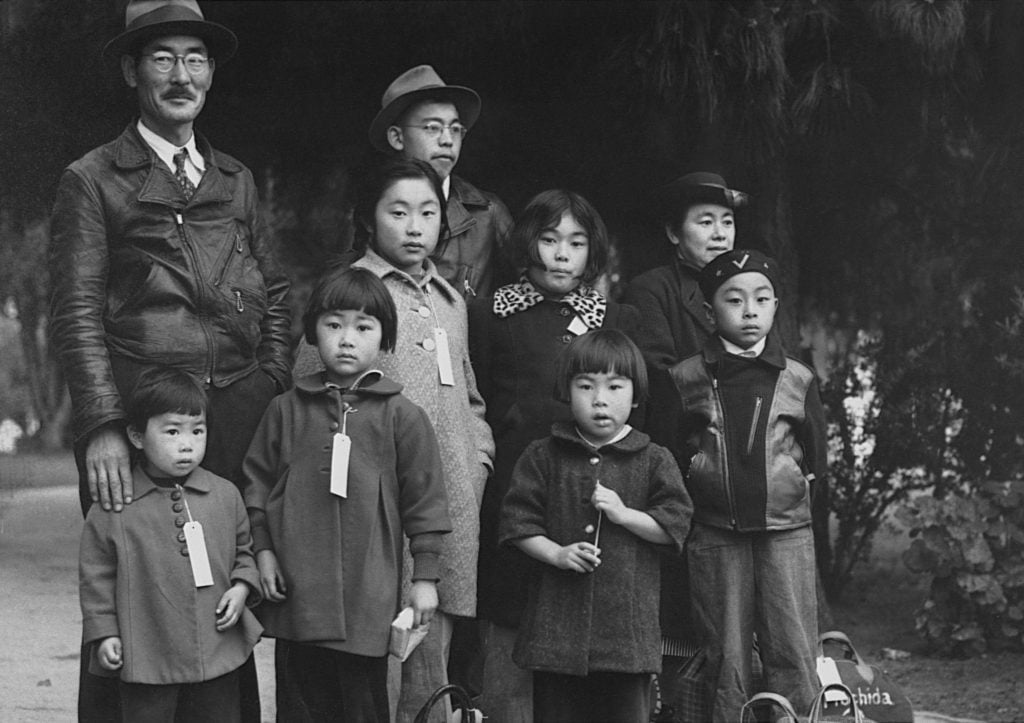 A black and white photograph of a family standing together in a cluster, wearing white tags that hang from their necks to their chests