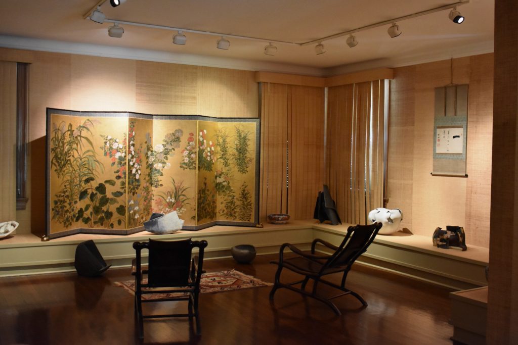 An elegant room of two chairs placed facing the wall, where a large, beautiful panel of Japanese art stands. 
