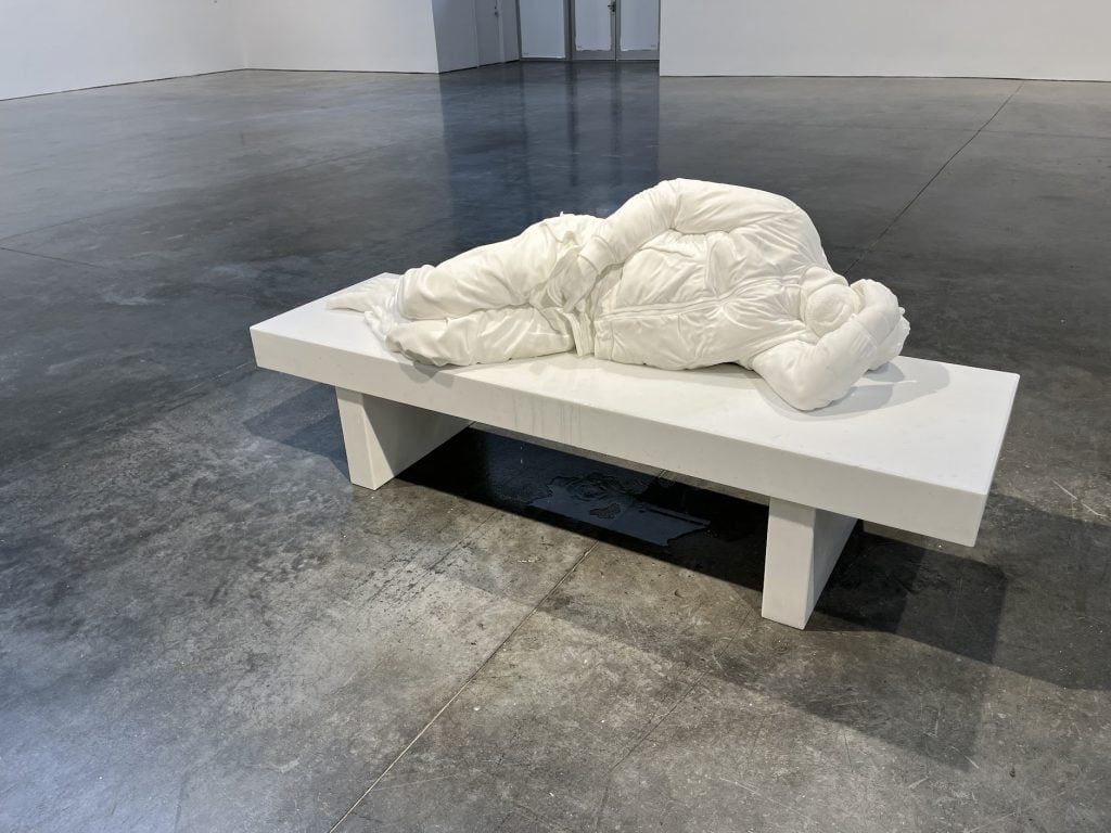 A color photo shows a white sculpture of a man lying on a bench in a cavernous space.