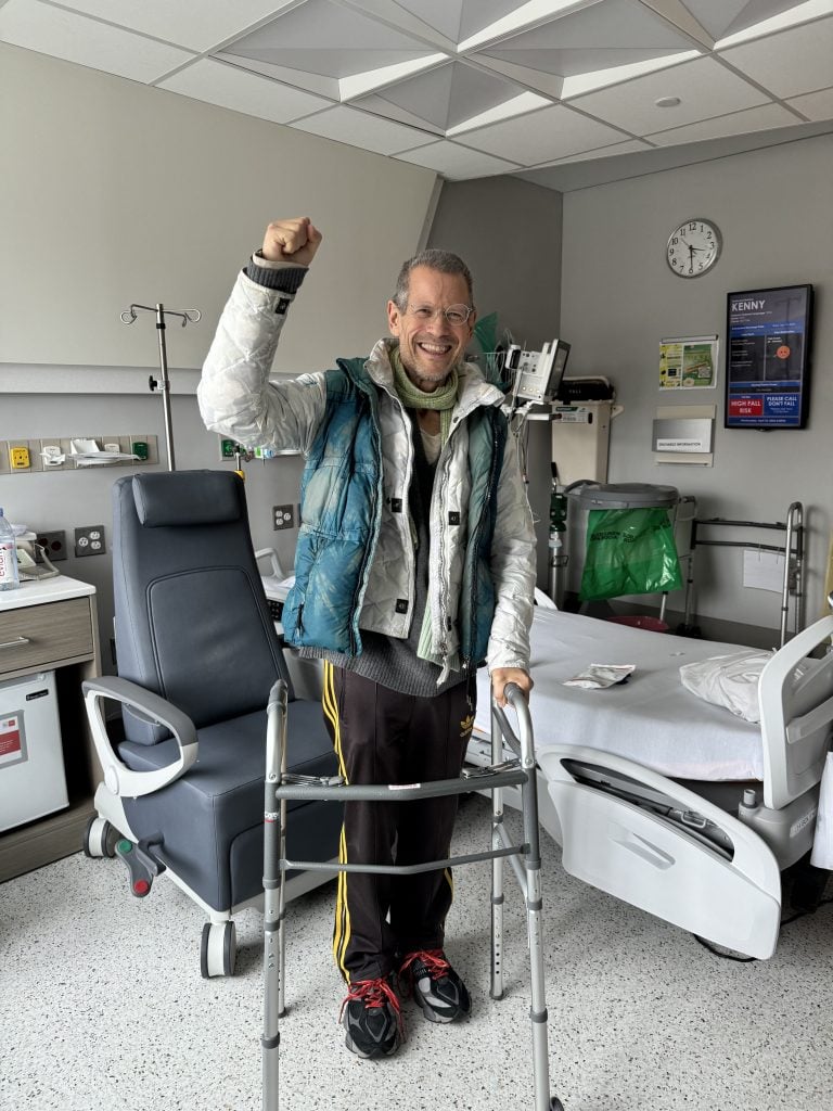 A man in casual clothes raises his fist in a color photo. He is in a hospital room, holding a walker.
