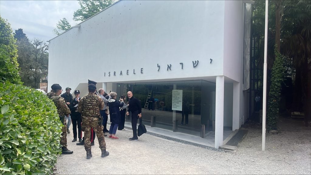 the image is of the israel pavilion at the venice biennale. the building is white and surrounded by trees. a crowd stands out front