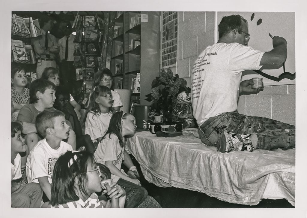 A group of captivated children sits on the floor of a classroom, watching intently as an artist paints a mural with whimsical black lines. The artist, focused on his work, is perched on a tarp-covered table, a jar of paint in one hand and a brush in the other. This educational setting is infused with a creative spirit, exposing young minds to the process of art-making in real-time.