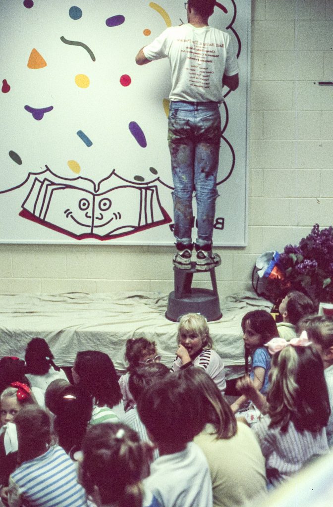 An artist stands on a stool, meticulously adding to a mural of cartoonish figures and shapes, as a group of children watch with fascination. The artist's clothing is splattered with paint, indicating a deep engagement with his creative work. The kids, some gazing up and others chatting, are part of an interactive art demonstration, learning firsthand about the artistic process.