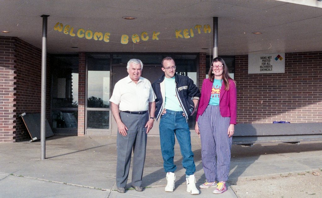 Three individuals stand smiling outside a building with a sign that reads "Welcome Back Keith," suggesting a warm greeting for someone named Keith. The older gentleman on the left is dressed in a classic white polo and gray slacks, while the young man in the center sports a retro, casual look with a letterman jacket, high-top sneakers, and acid-wash jeans. The young woman on the right wears a vibrant ensemble featuring a checkered pair of pants and a colorful sweater, embodying the lively fashion sense of a past era.