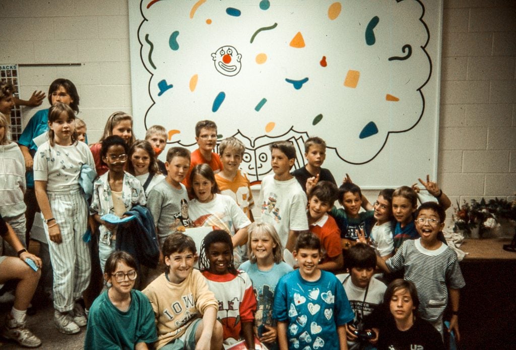 A group of cheerful children pose for a photo in front of a playful mural, filled with whimsical shapes and a cartoon-like cloud. Their varied expressions and casual 90s attire add to the nostalgic charm of the image. The mural's simple, joyful design serves as a fitting backdrop for the youthful energy of the group.