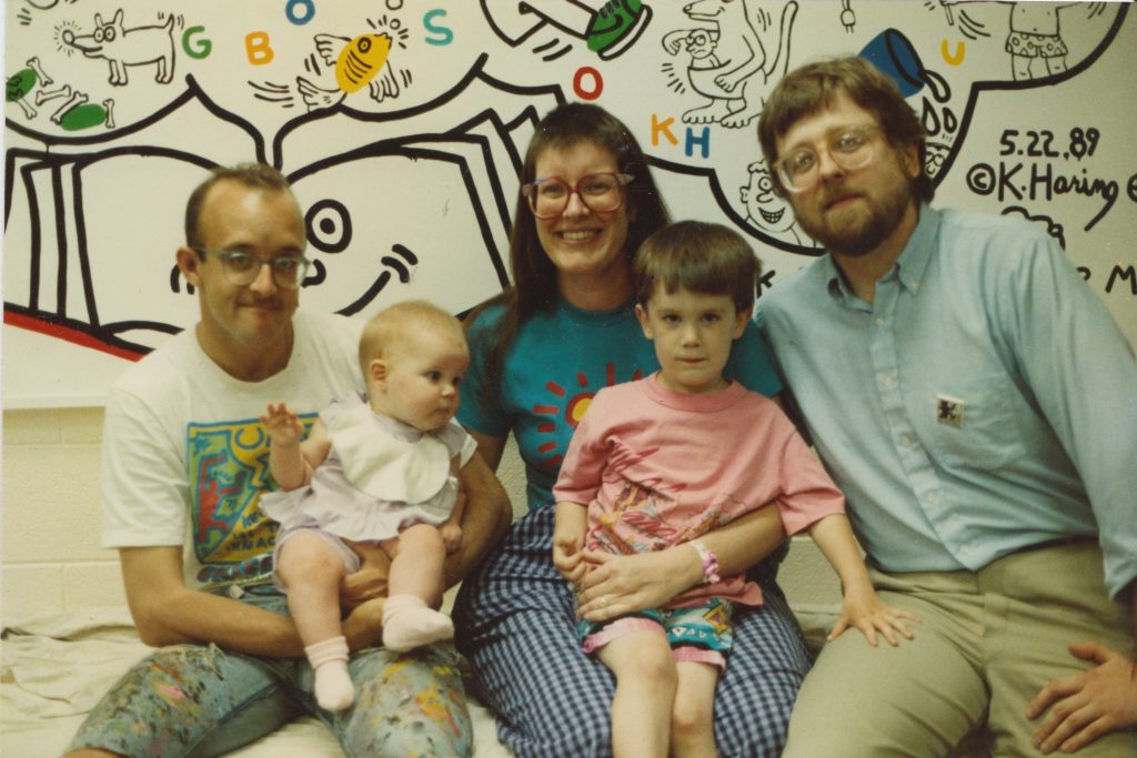A family sits together in front of a playful, pop-art style mural by Keith Haring, who holds a baby in the left of the frame. Also pictured are a woman with glasses, alongside a child in pink, smiling for the camera with another man.