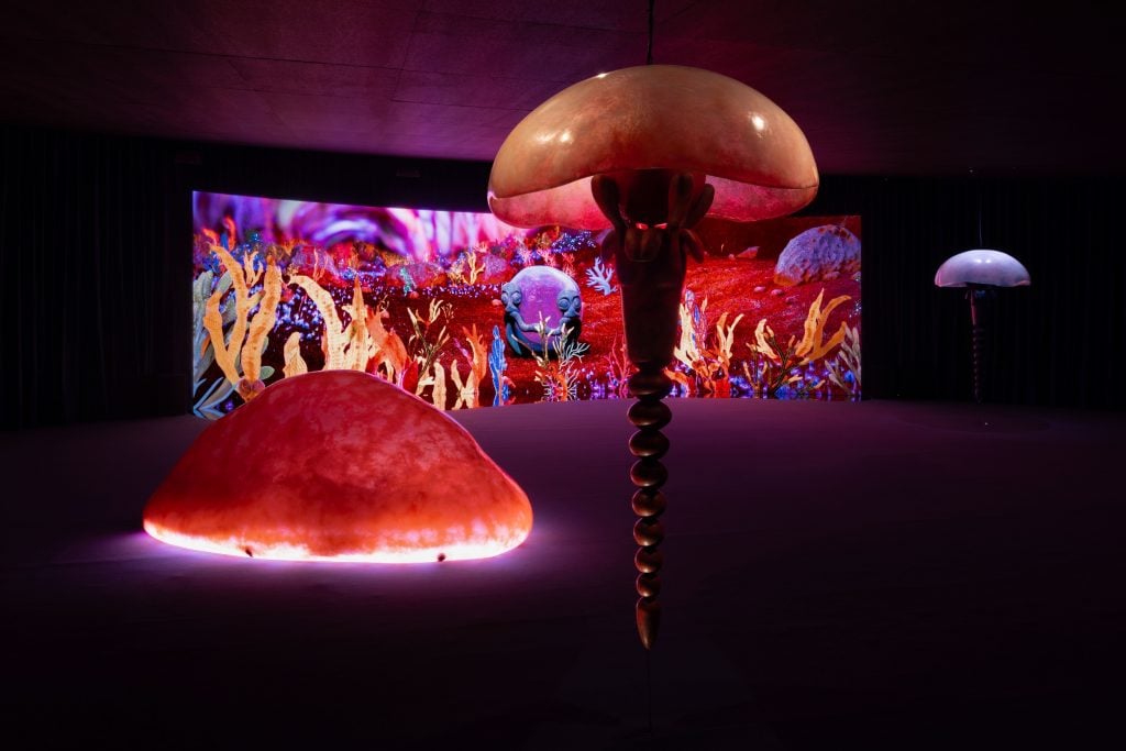 large glowing red resin sculptures of jelly fish hover in front of a screen showing a psychadelic, colorful scene reminiscent of the sea floor