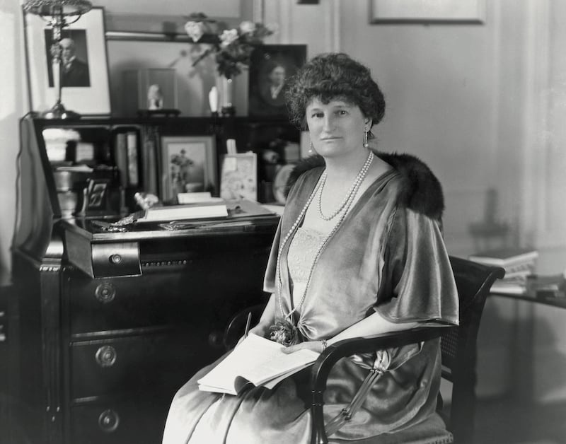 A 1922 photograph of a woman sitting at a desk, an open book in her lap, and a satin robe across her shoulders.