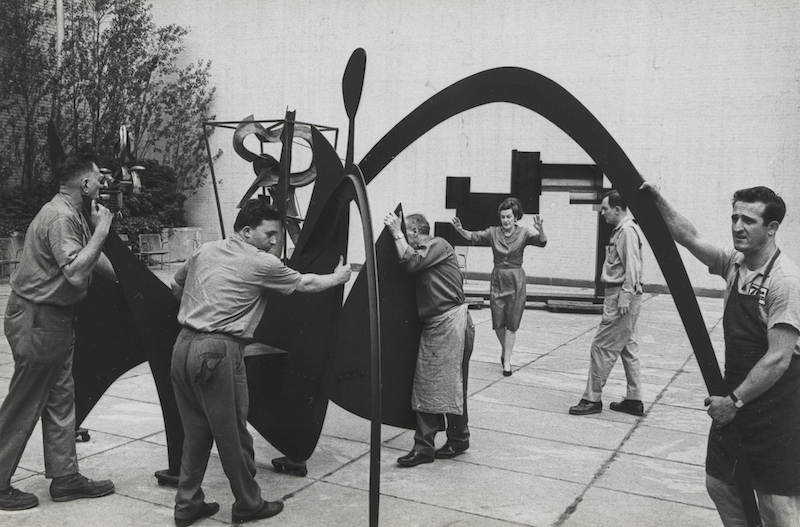 A black and white photo of a team stationing a sculpture while a woman gesticulates, providing energetic guidance