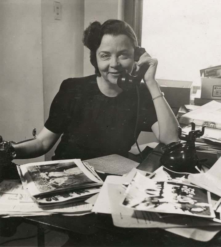 A black and white photograph of a women seated and posed behind a desk laid out with pages of images