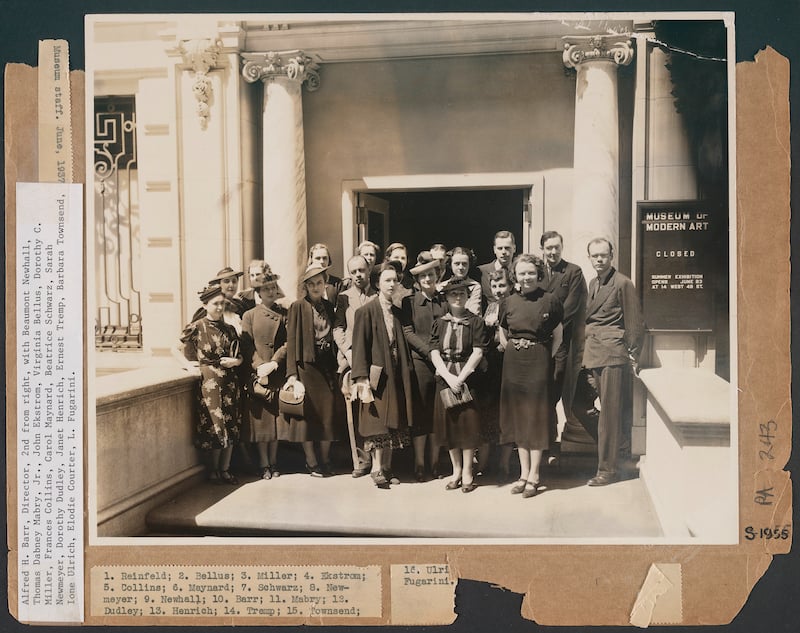 A sepia-tinged old photo of a professionall-dressed group of MoMA employees from Inventing The Modern posing for a group photo outside, between two grand columns