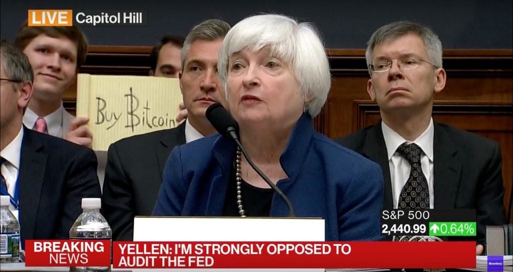 A woman sitting at a desk during a congressional hearing. Behind her, a man holds up a legal pad that reads "Buy Bitcoin."