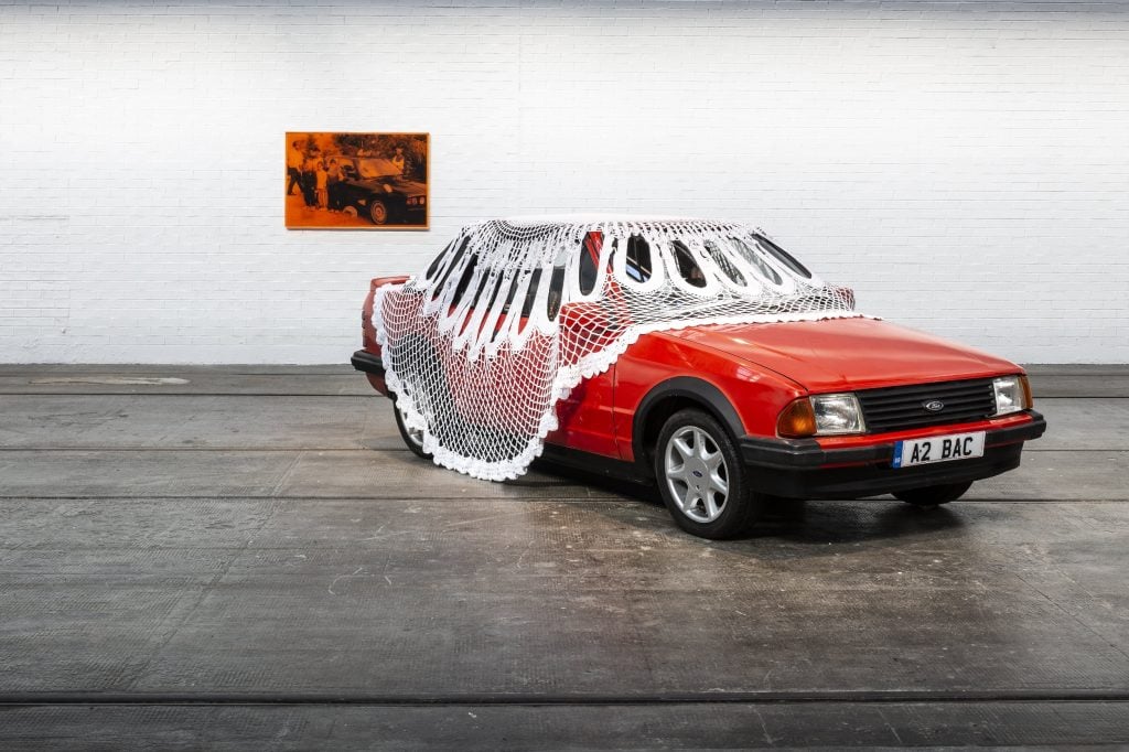 a red vintage Ford Escort covered in a very large doilly with a faded photograph of the car hanging on the wall behind