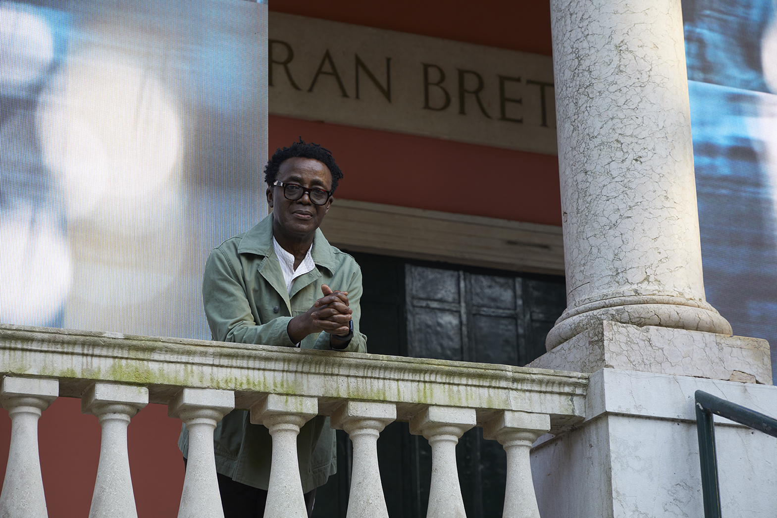 A man with glasses, the artist John Akomfrah, leans over a mossy balcony.