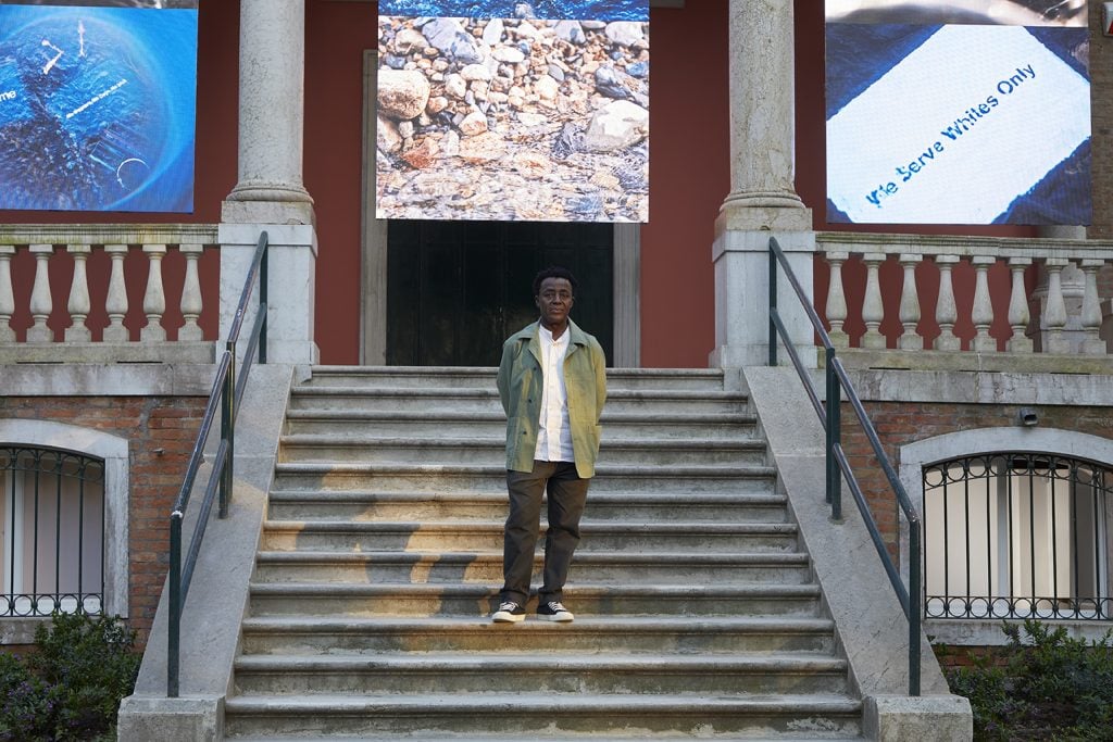 artist John Akomfrah stands on the stairs of a large building with videos playing behind him