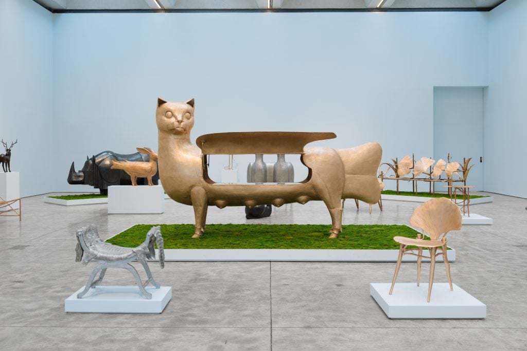 A large gold sphinx-like cat sculpture is in the foreground of a gallery standing atop fake grass surrounded by sculptures that blend animals and plants into furniture 