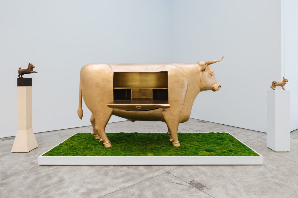 A large gold Les Lalanne statue of a cow sits atop fake grass, it doubles as a cabinet
