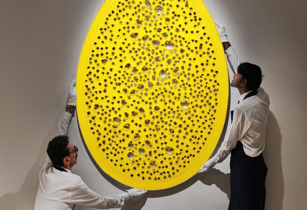 Two men in white gloves position a large yellow oval-shaped painting by artist Lucio Fontana on a white gallery wall.