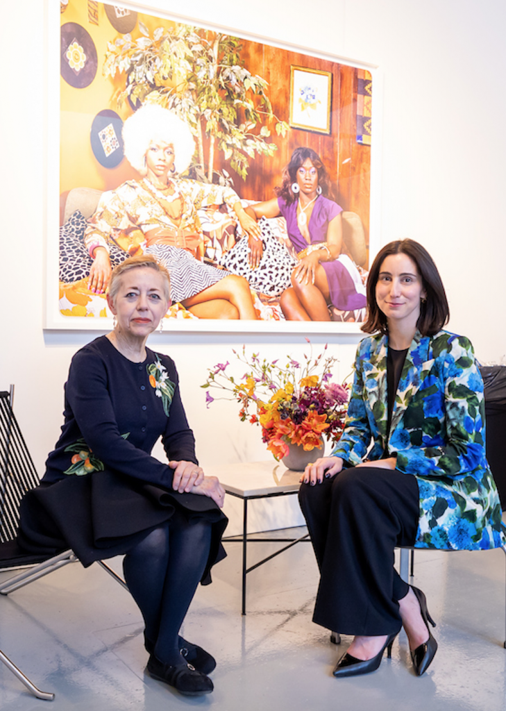 two white women look out of the picture, they are seated at a small table with flowers in an office like setting, both are dressed professionally, on the wall behind them is a photograph of two black women on a couch in what appears to be a 1970s intertior