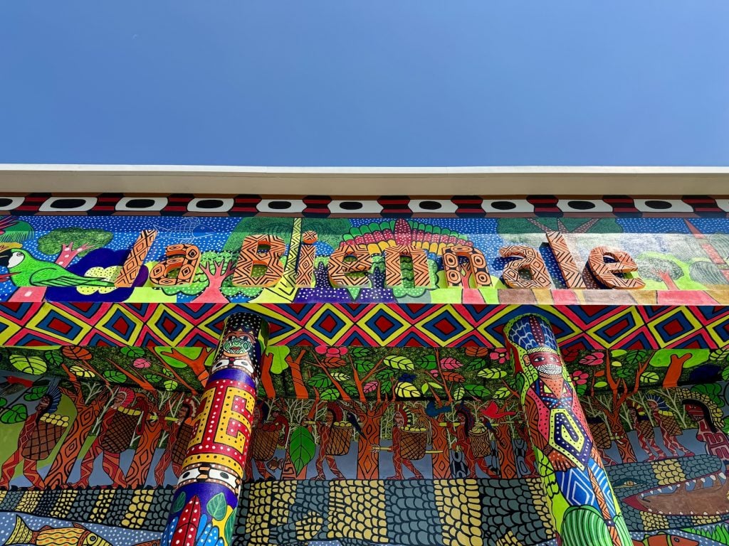 The front of a building viewed from below that says 'a Biennale,' covered with brightly colored patterns