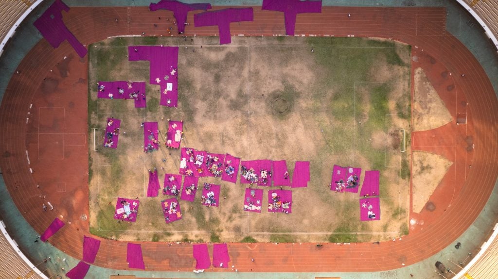a drone aerial image shows a standium's central grass patch filled with large sections of bright pink material and people can be seen dotted around working on the material