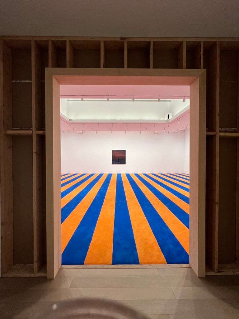 A room with an orange and blue striped carpet on the floor and a dark painting on the back wall