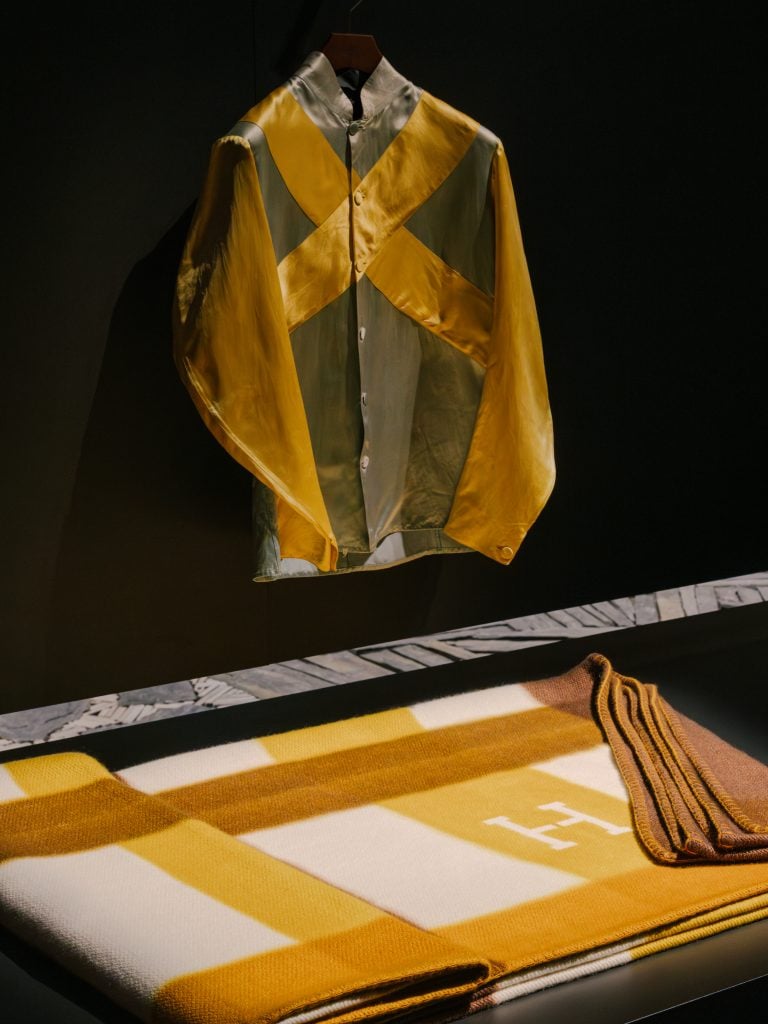 a green and yellow jockey shirt is positioned above a brown and yellow plaid blanket in a showcase by Hermès
