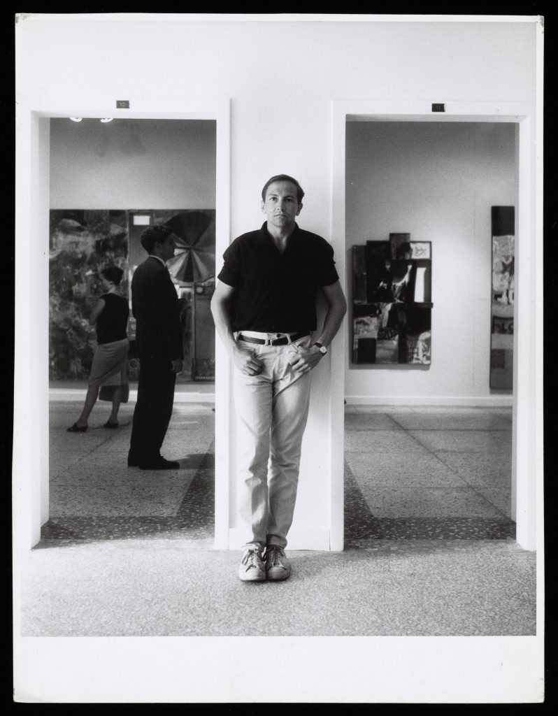A man, artist Robert Rauschenberg, standing between two doorways leading to galleries hanging with art