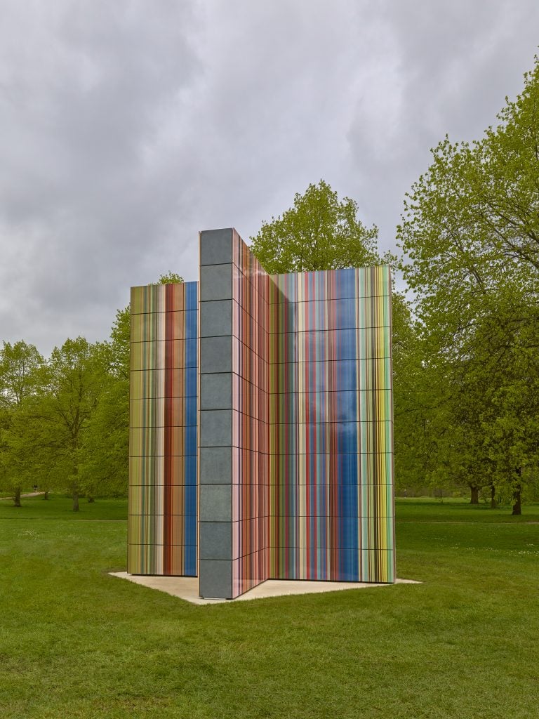 a geometric sculpture with lines of color stands in the middle of a grassy park
