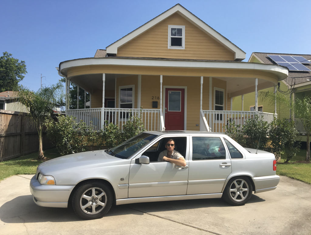 A man leans out of a silver car in front of a yellow house.