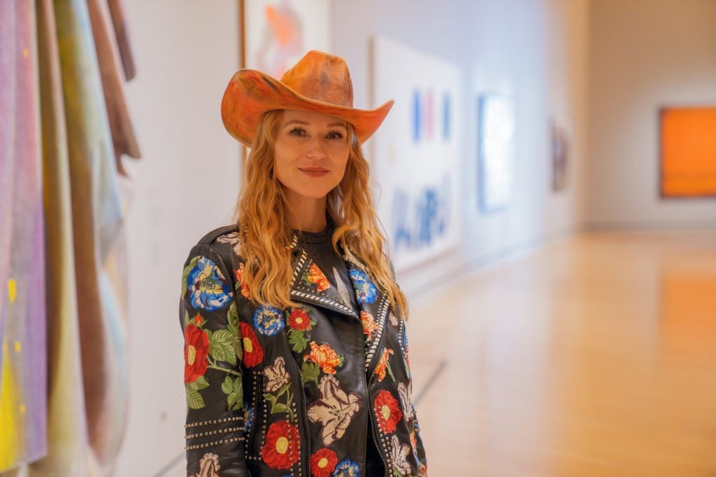 a woman in a cowboy hat stands in a museum. there are artworks hanging on the all behind her. she is smiling at the camera