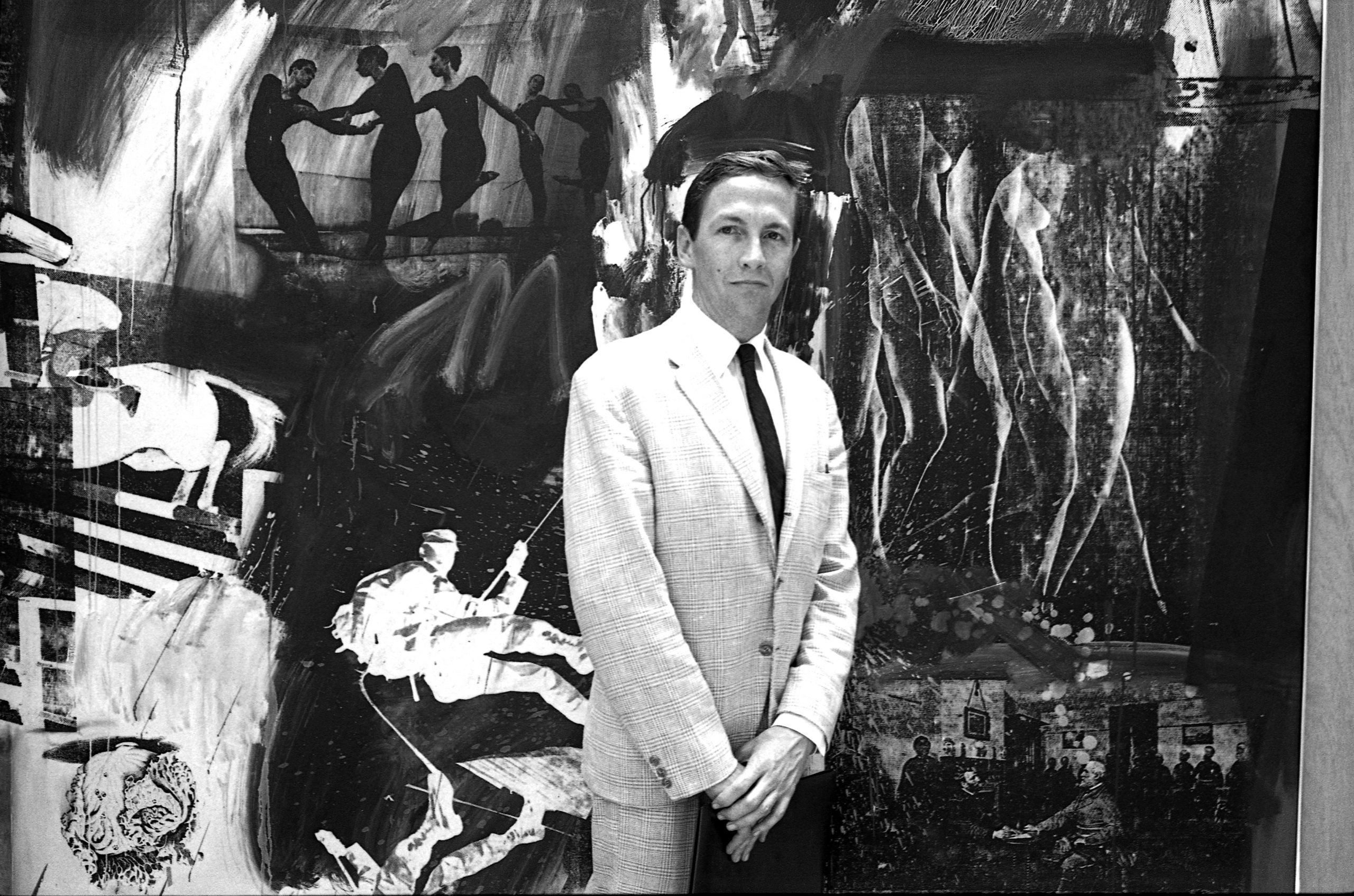 A black and white photo of a man in a white suit, artist Robert Rauschenberg, standing in front of an abstract canvas