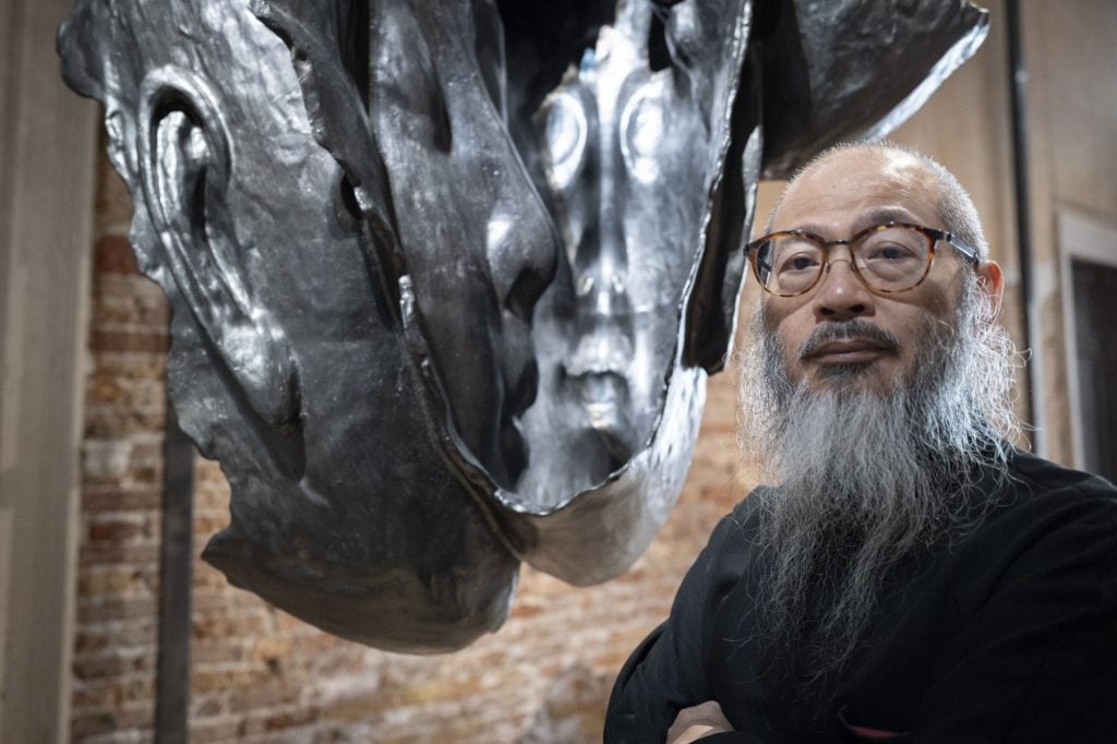 A man with a long beard posing next to a dark coloured sculpture