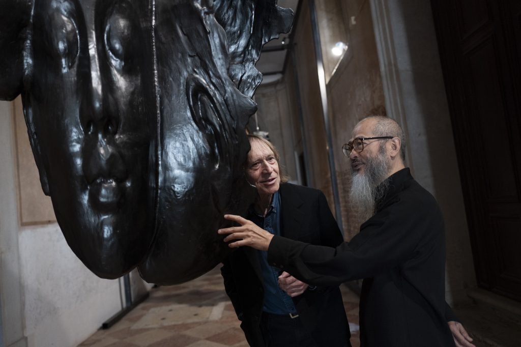 A man with a long beard looking at a big sculpture, next to a caucasian man. 