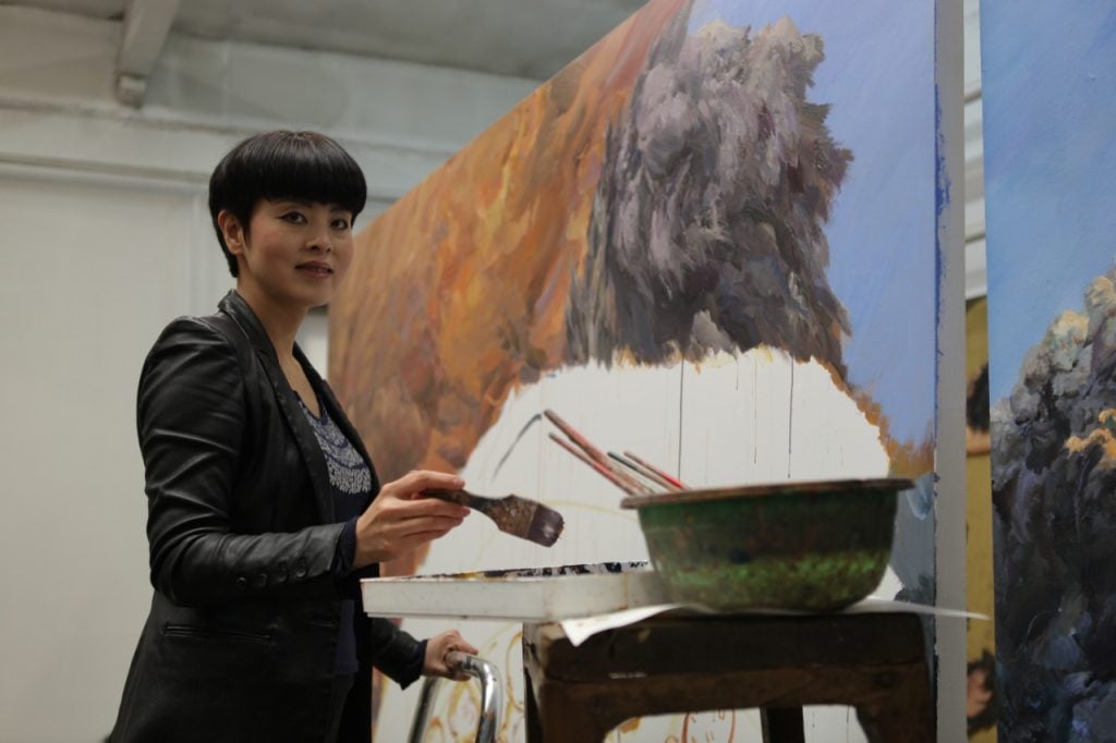 An asian woman stands in front of a massive canvas, mid-paint