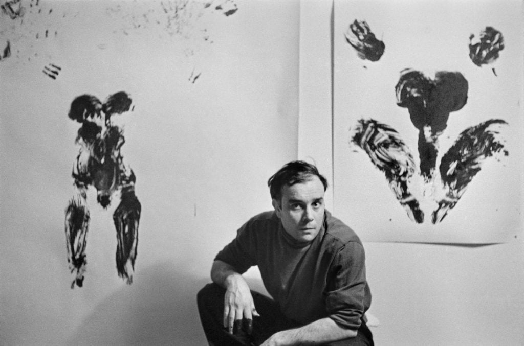 the artist Yves Klein squats in front of two of his works, imprints of female bodies on paper