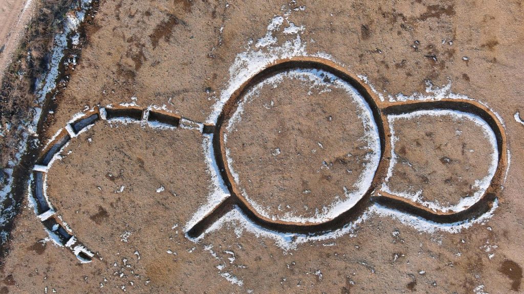An aerial photograph of a circle indentation in dirt, with a petal-like appendage and another shaped like an open horseshoe