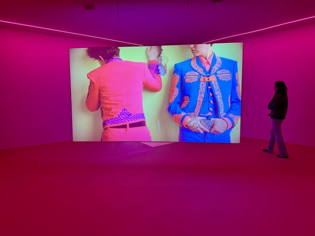 A screen depicting two men in brightly colored outfits slapping one another in a bright pink gallery