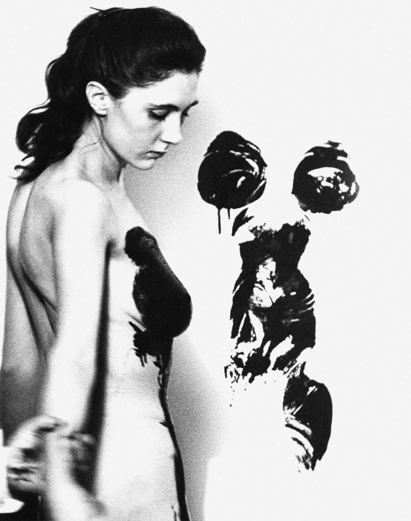 A naked woman with paint on her body has pressed herself against a canvas, leaving a mark in her shape